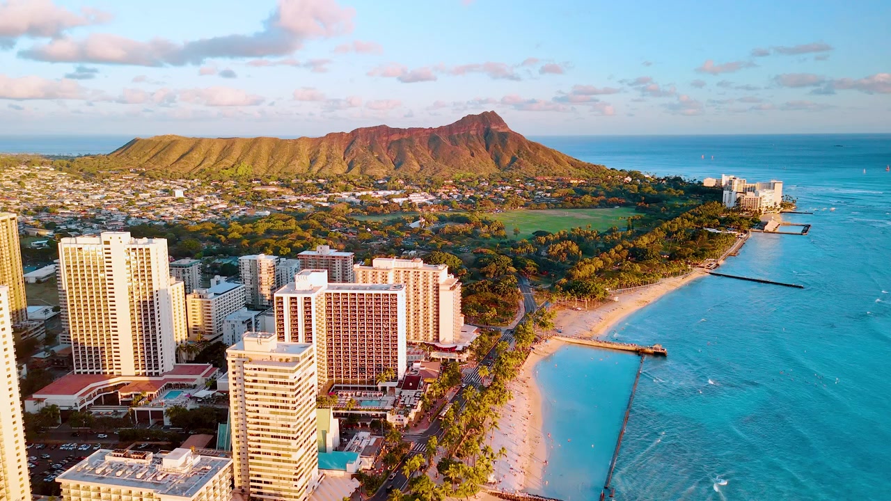 Hotel della spiaggia di Waikiki e Diamond Head da un'angolazione elevata lato oceano, Hawaii. Fila di torri alberghiere accanto alla costa sabbiosa, molo frangiflutti roccioso, acqua turchese, cratere vulcanico e oceano aperto all'ora d'oro.