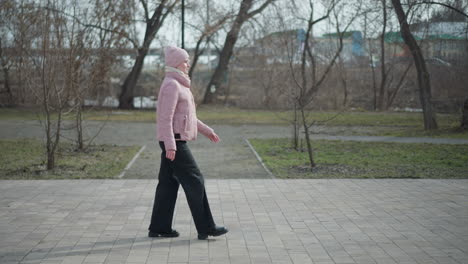 Young woman in pink jacket and beanie walks through park during cold season, partially obscured by vertical black pole, surrounded by leafless trees, gray pavement, and subdued outdoor atmosphere