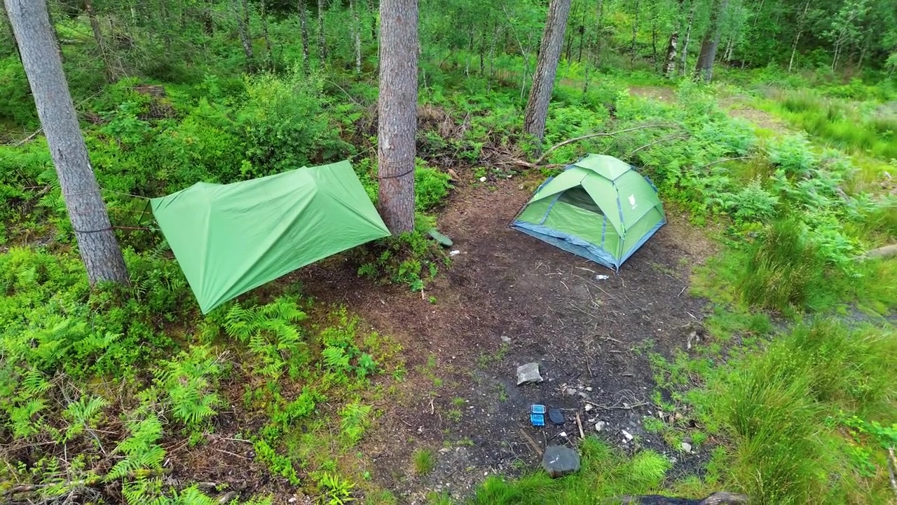 Acampamento na floresta visto de cima enquanto um drone gira lentamente para a direita, revelando uma barraca cúpula verde e um abrigo de lona ao lado de pinheiros, samambaias e um pequeno círculo de fogo em uma área limpa