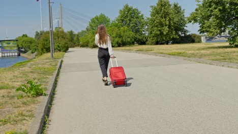 Female-tourist-walking-with-red-suitcase-over-empty-road-along-river,-Riga