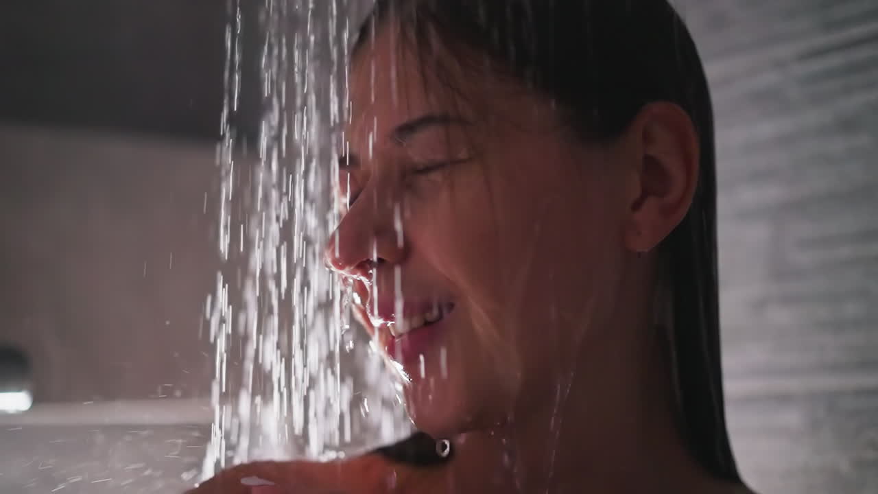 샤워실에서 물 스프레이에 미소 짓는 여자. 눈을 감은 쾌적한 갈색 여자가 화장실에서 몸을 고 휴식을 취합니다. 집에서 레크리에이션 시간