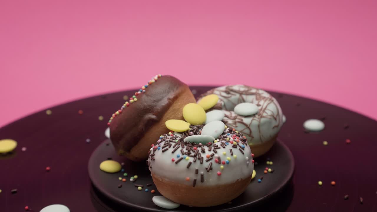Three American donuts spin in circles on a black and pink surface. They are topped with black and blue chocolate and choco and colorful crumbs