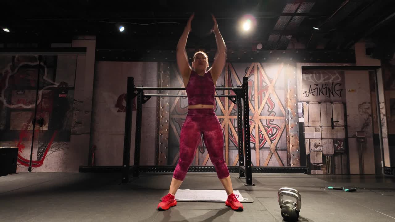 A fit woman in a gym performs a functional ground to overhead exercise with a medicine ball, showing strength and power during her full-body workout