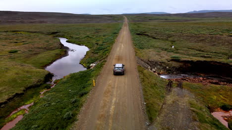 Luftfahrt-Eines-Autos-Von-Hinten-Auf-Einer-Einsamen-Bergschotterstraße,-Umgeben-Von-Grünen-Büschen-In-Island