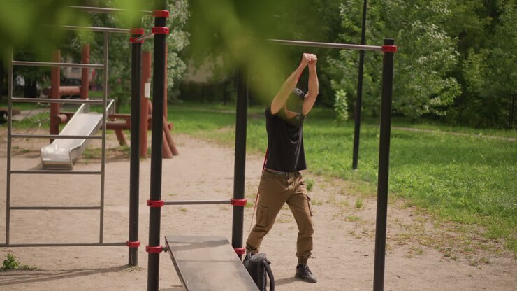 Park Overhead Mobility And Shoulder Stabilization Sequence With White Male Clasping Hands Above Head And Moving Under Bar, Focused Breathing, Cargo Pants And Casual Cap, Natural Green Background