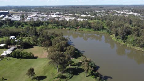 4k drone shot approaching a brown river flying over a green park, showing an industrial estate on the other side of the river in australia