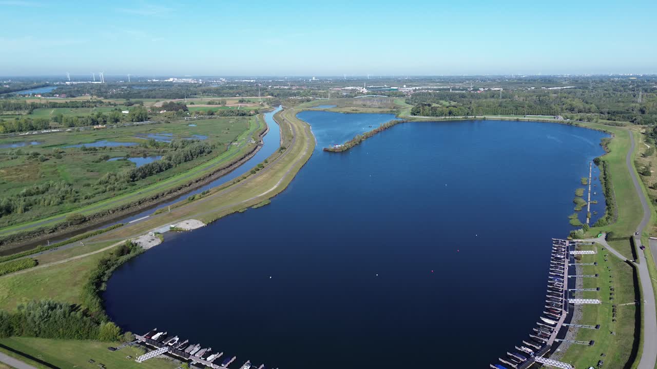 Drone flies north over the lake. Piers with boats, the Dijle River and lush nature are visible. In the distance, several wind turbines rise above the landscape near Mechelen, Belgium