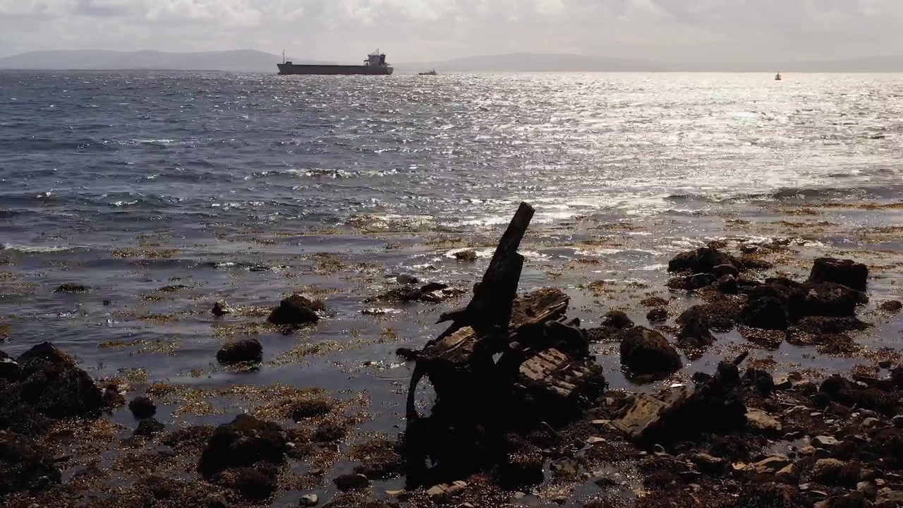 Vista estática de costa rochosa com barco de madeira quebrado na margem, uma barcaça de transporte ao longe, destacando o cenário costeiro da Ilha Mutton.