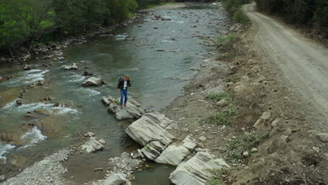 Male-hiker-walking-through-rocky-river-shore-jumping-on-rocks-among-green-trees