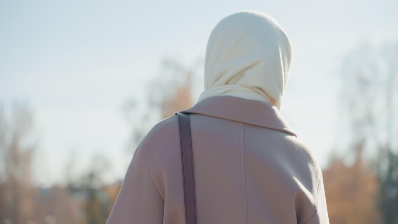 Back view woman wearing headscarf and wool coat in sunlit autumn park, soft bokeh of orange trees, shoulder bag on strap, slow contemplative pause, cool pastel tones and gentle breeze