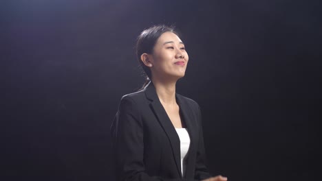 블랙 스크린 스튜디오에 서서 손을 치고 주위를 둘러보는 미소 짓는 아시아 스피커 여성