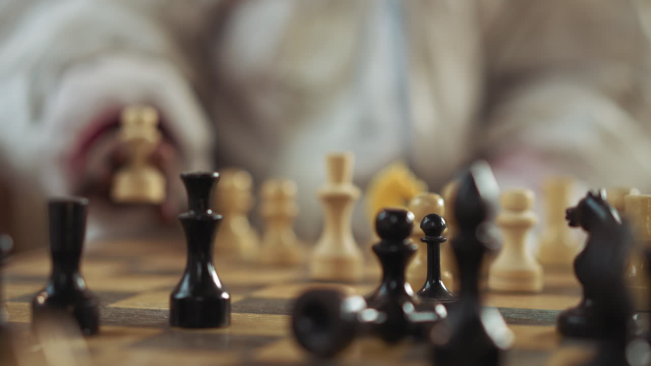 Close up of hand moving white chess piece on wooden board surrounded by black and white pieces during indoor match, showing strategic tension with blurred figure in protective clothing in background