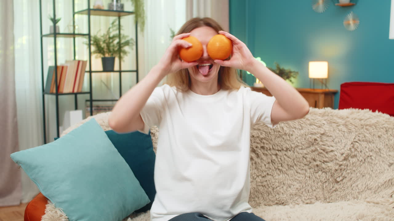 Pregnant woman makes funny faces with oranges over eyes laughs fresh vitamin fruit fun at home sofa