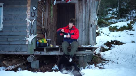 lone man in a wooden chalet eating outside with dog pet in winter countryside