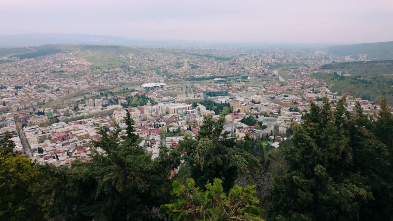 Rozsáhlý pohled z výšky na panorama Tbilisi v Gruzii, s hustou městskou zástavbou, historickými budovami a moderními stavbami orámovanými svěžími zelenými stromy v popředí. Opar