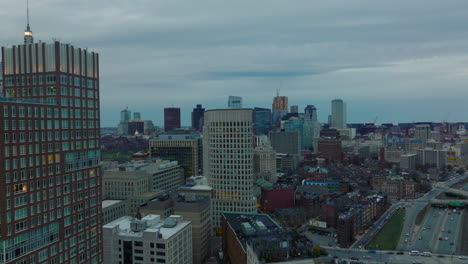 Backwards-reveal-of-buildings-in-wider-downtown.-Aerial-view-of-high-rise-office-buildings-and-busy-highway-at-dusk.-Boston,-USA