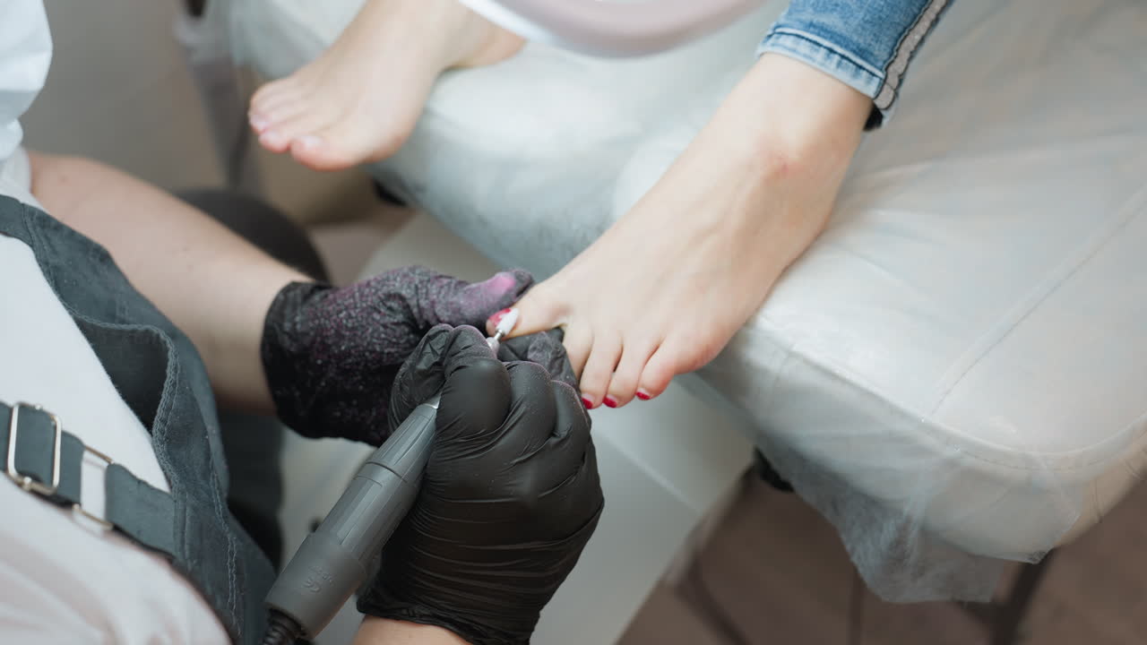 Upward view of nail technician filing client toenail using electric tool while client relaxes on padded chair, lamp positioned above for enhanced visibility during precise foot grooming session