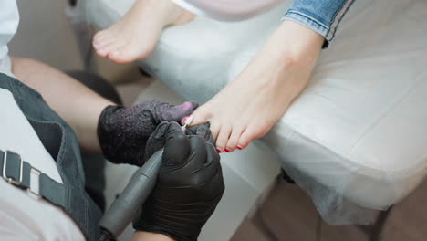 upward view of nail technician filing client toenail using electric tool while client relaxes on padded chair, lamp positioned above for enhanced visibility during precise foot grooming session