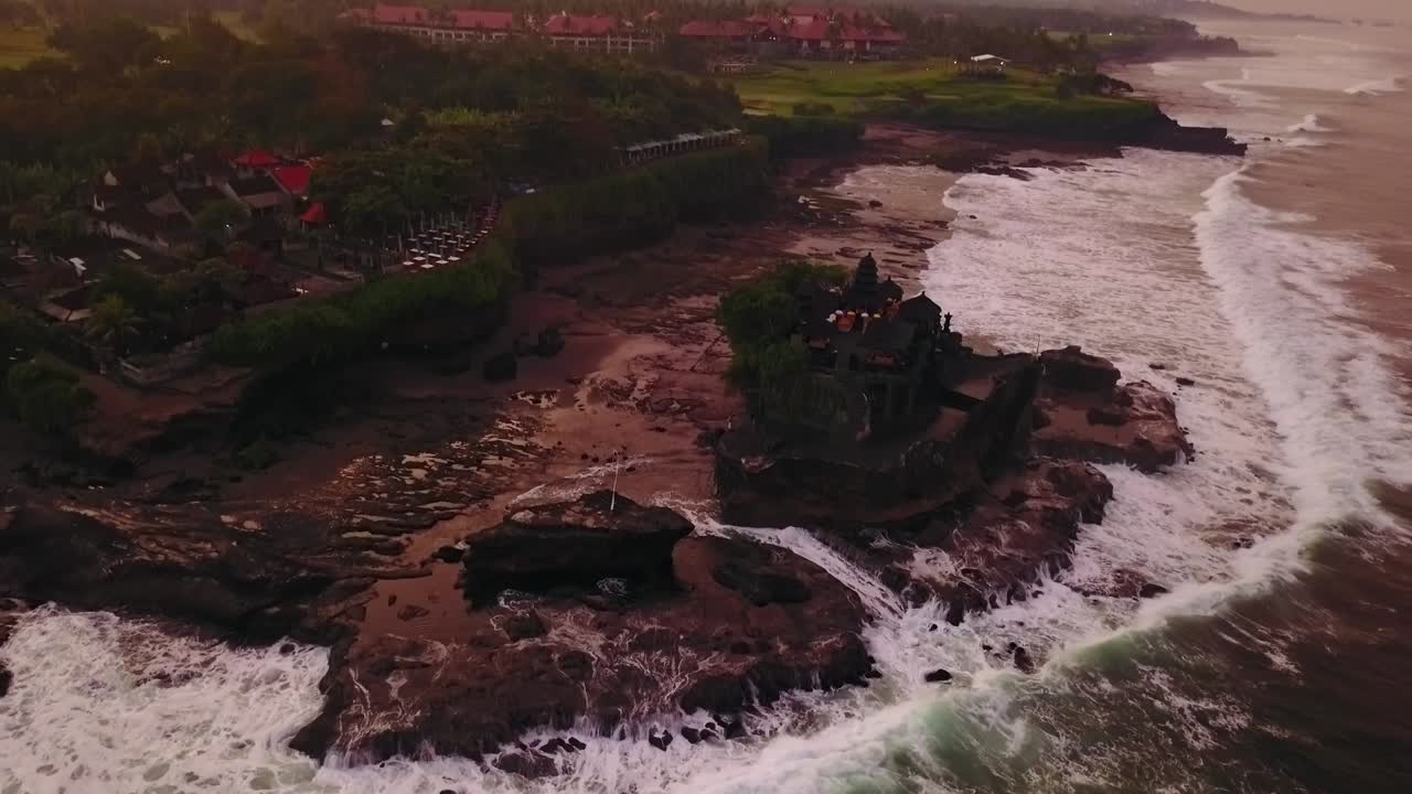 Aerial view of Tanah Lot temple and coastline at sunset. Captures the iconic temple against the backdrop of the vibrant sky and rugged shore.
