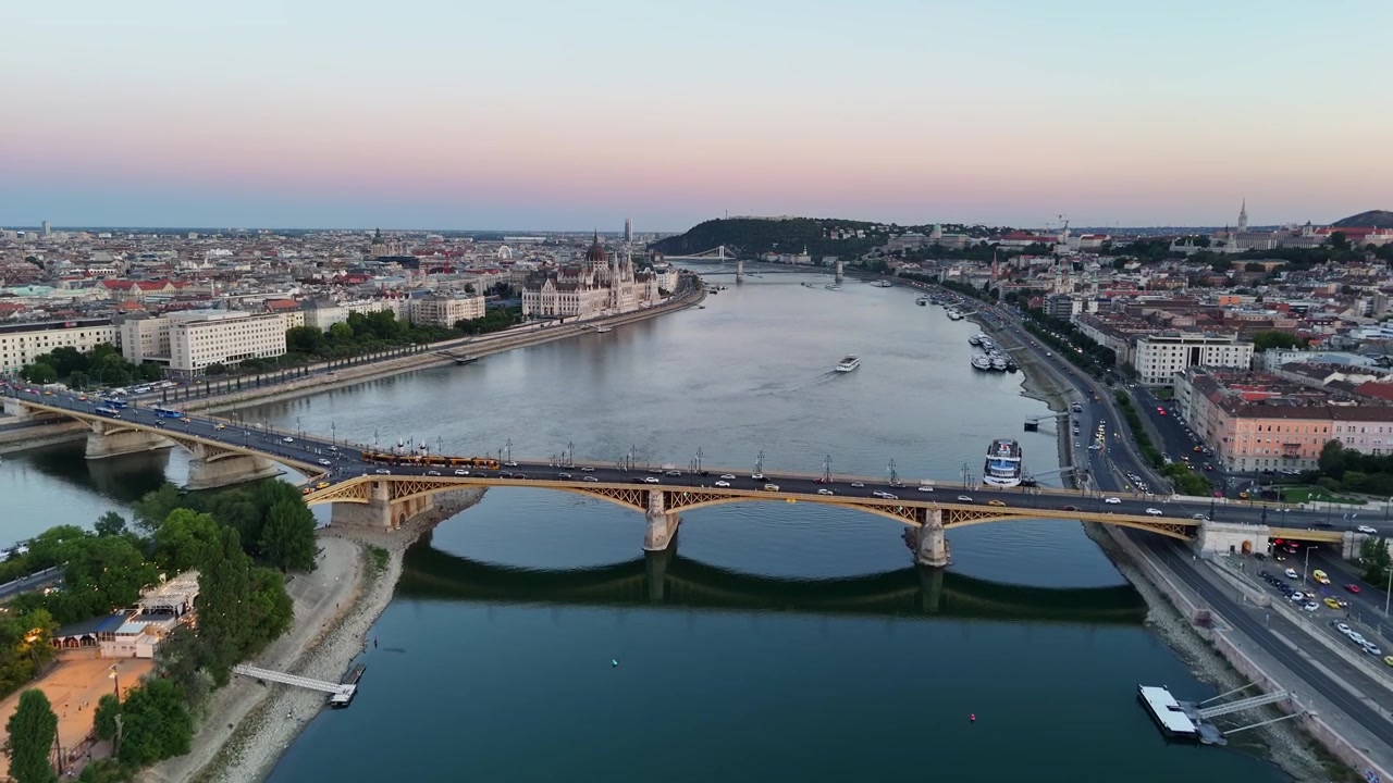 Budapest, Ungheria: il Parlamento ungherese e il Ponte Margherita lungo il Danubio al crepuscolo, con bassi livelli dell'acqua, barche, traffico e linee del tram visibili in una ripresa aerea con drone