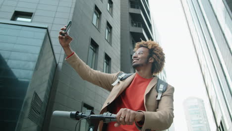 positive african american man standing in city with e-scooter and taking selfie