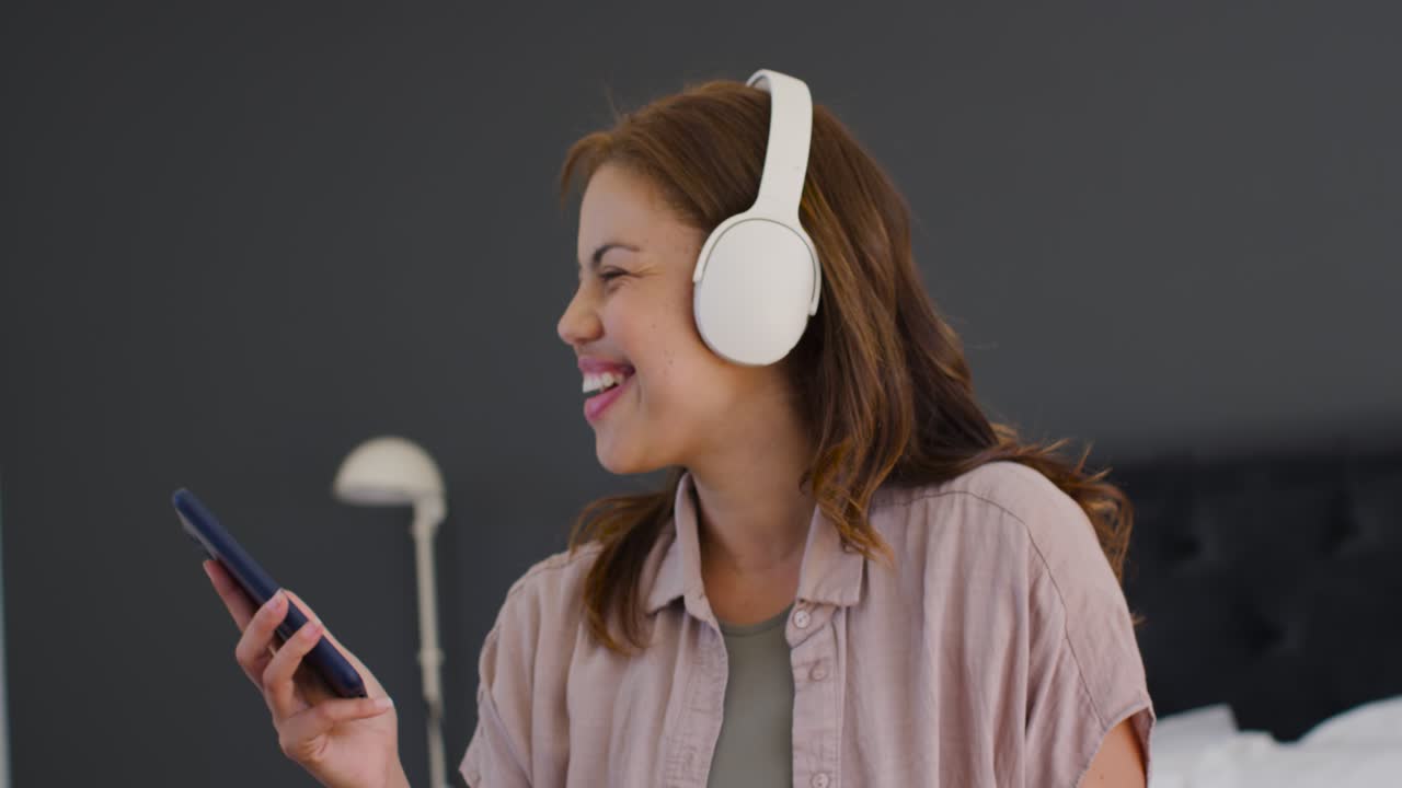 Donna afroamericana con telefono in mano, che indossa cuffie bianche, ascolta musica e canta sul letto.