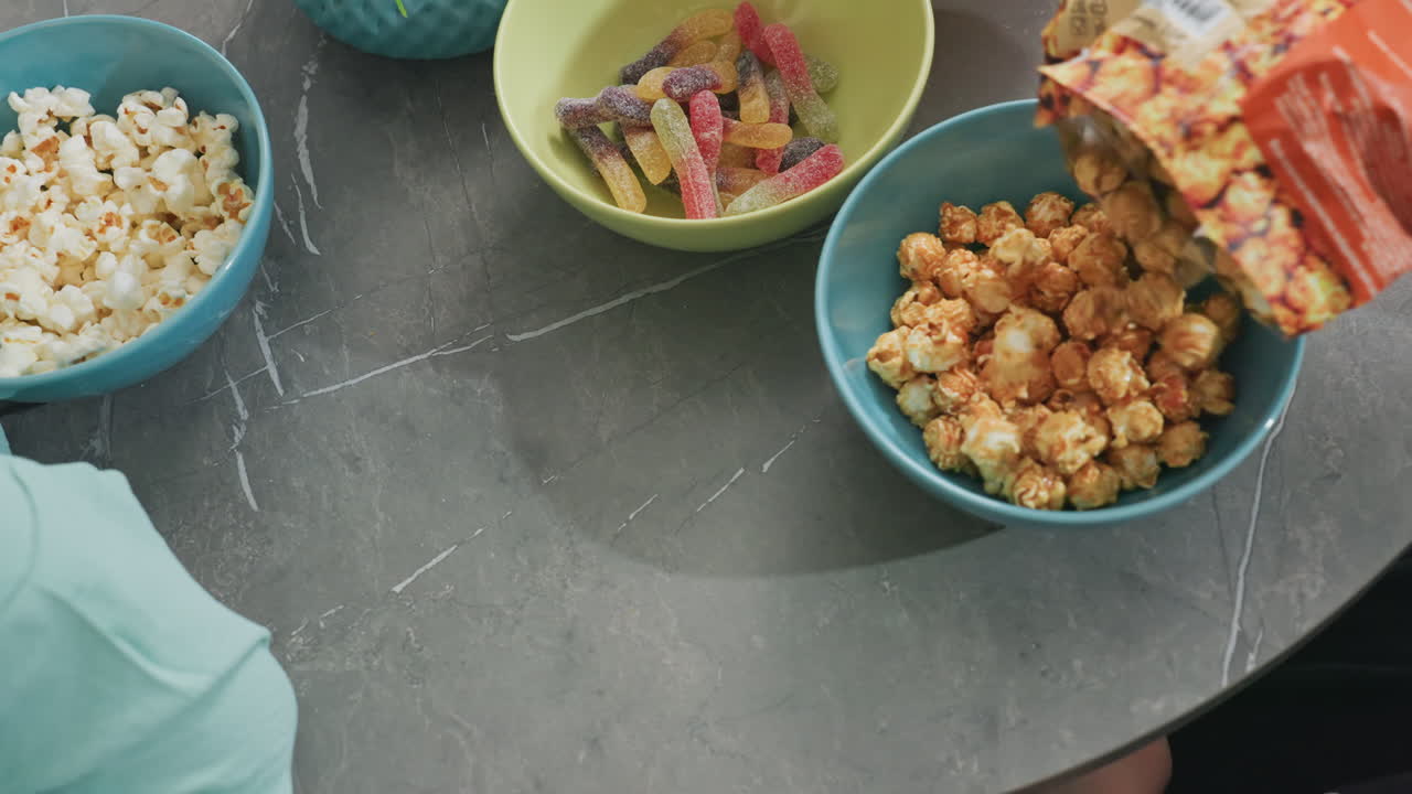 top down view of person pouring flavored popcorn into bowl on kitchen table with bowls of gummy candy and chips nearby, casual snack time, home setting, colorful bowls, food preparation