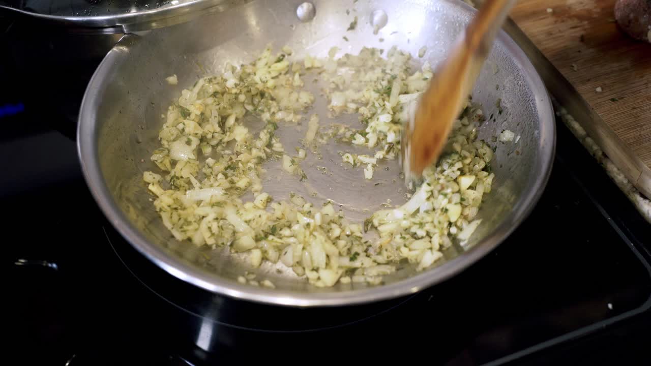 Using wooden spoon to turn onions and thyme inside of steel pan Preparing ingredients to make vegan beyond meatballs with spaghetti and meat sauce