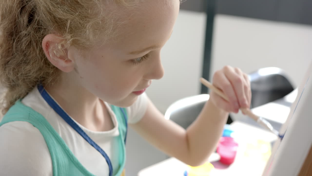 A girl with blonde hair is painting, focused on her artwork, in a classroom at school