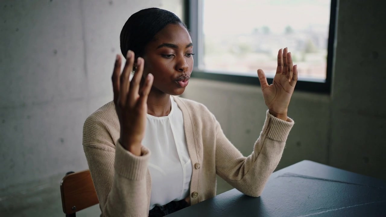 Starting speech, presenter wearing beige cardigan, white blouse gesturing at dark table, window