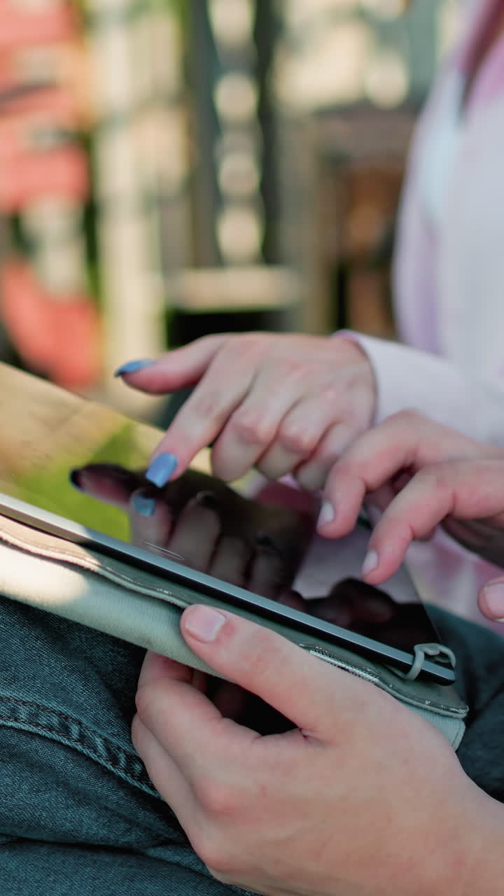 primer plano de una mujer con una camiseta rosa y uñas pulidas operando una tableta mientras continúa escribiendo en su computadora portátil, sentada al aire libre con un hombre escriviendo en su propio dispositivo