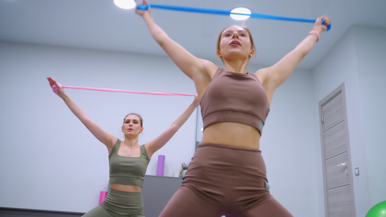 두 명의 여성이 잘 조명 된 체육관에서 다채로운 저항 리본으로 역동적으로 스트레칭하여 체력과 유연성을 보여주고 있습니다.