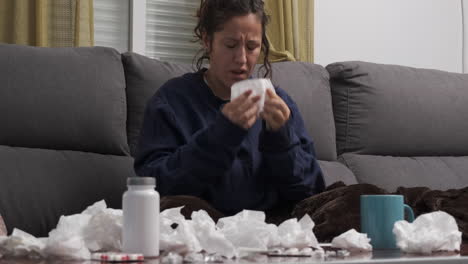 young woman sick on the sofa at home, blowing her nose with handkerchiefs. pills, tissues and a cup on the table