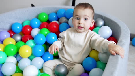 adorable toddler sits in a dry pool. cute boy leans back and lies in the multiple balls smiling to the camera.