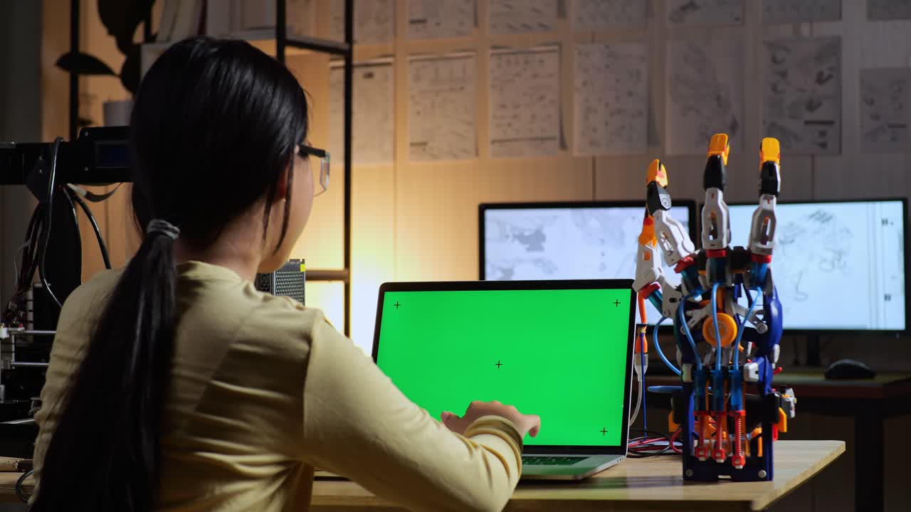 십대 아시아 소녀가 집에서 녹색 화면 모형을 가진 노트북에서 사이보그 손을 설계하는 뒷면