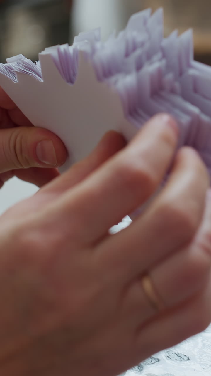 Unas manos caucásicas doblan un copo de nieve de papel con cuidado sobre una mesa de cristal, apilando y doblando hojas de color lila pálido para montar una guirnalda. Se ve un anillo de boda y un suave movimiento táctil.