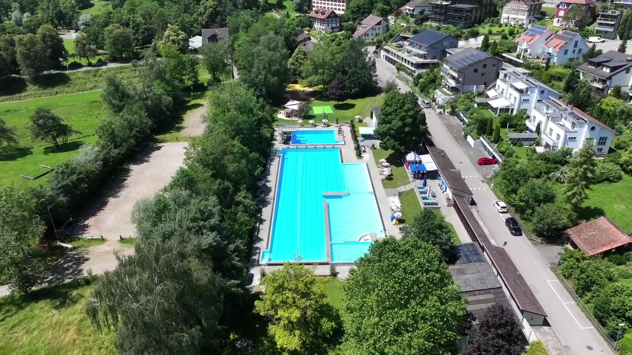 Luftfoto med udsigt over en udendørs klar blå swimmingpool i et naturskønt schweizisk byresort
