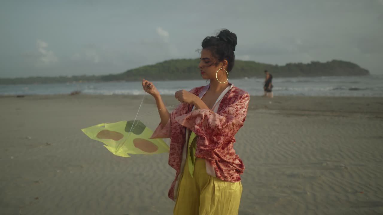 A young Asian adult, donning a contemporary attire and showcasing a distinctive hairstyle, stands on a breezy beach, grappling with the challenge of flying her kite