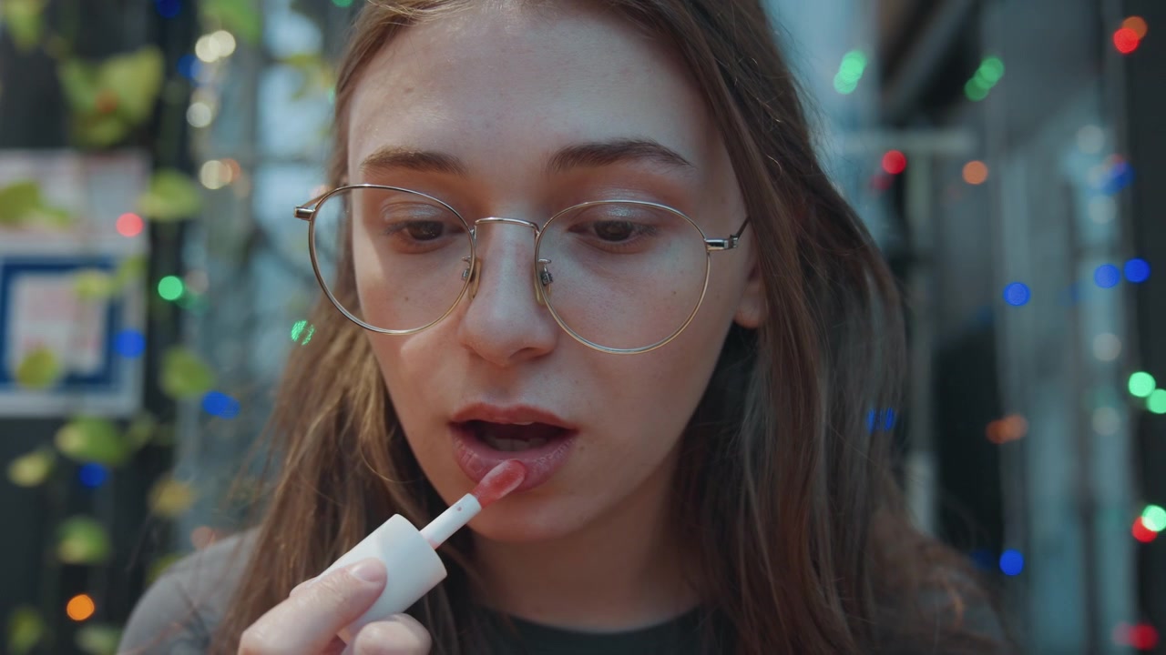 Youthful woman applying lipgloss. Candid shot of woman with glasses. Candid moment capturing young woman wearing glasses applying lip balm in warm cafe setting with swirling bokeh lights