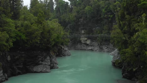 Aerial-of-swing-bridge,-rocks,-native-forest-at-Hokitika-Gorge,-New-Zealand-on-a-rainy-day