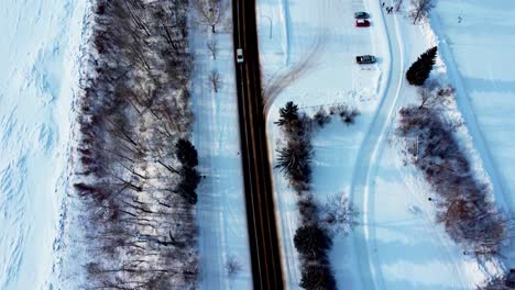 Vista-Aérea-De-Pájaros-De-Invierno-2-Carretera-De-Dos-Carriles-Junto-Al-Río-Del-Parque-Cubierto-De-Nieve-Y-Pista-De-Patinaje-Vacía-Y-Tranquila-Y-Pista-De-Hielo-Con-Arbustos-Y-árboles-A-Su-Alrededor-Y-Algunos-Autos-Estacionados-Siguiendo-A-La-Camioneta-Blanca-3-3