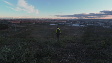 vista trasera del hombre con chaqueta amarilla y mochila caminando cuesta abajo con el horizonte de madrid en la distancia temprano en la mañana