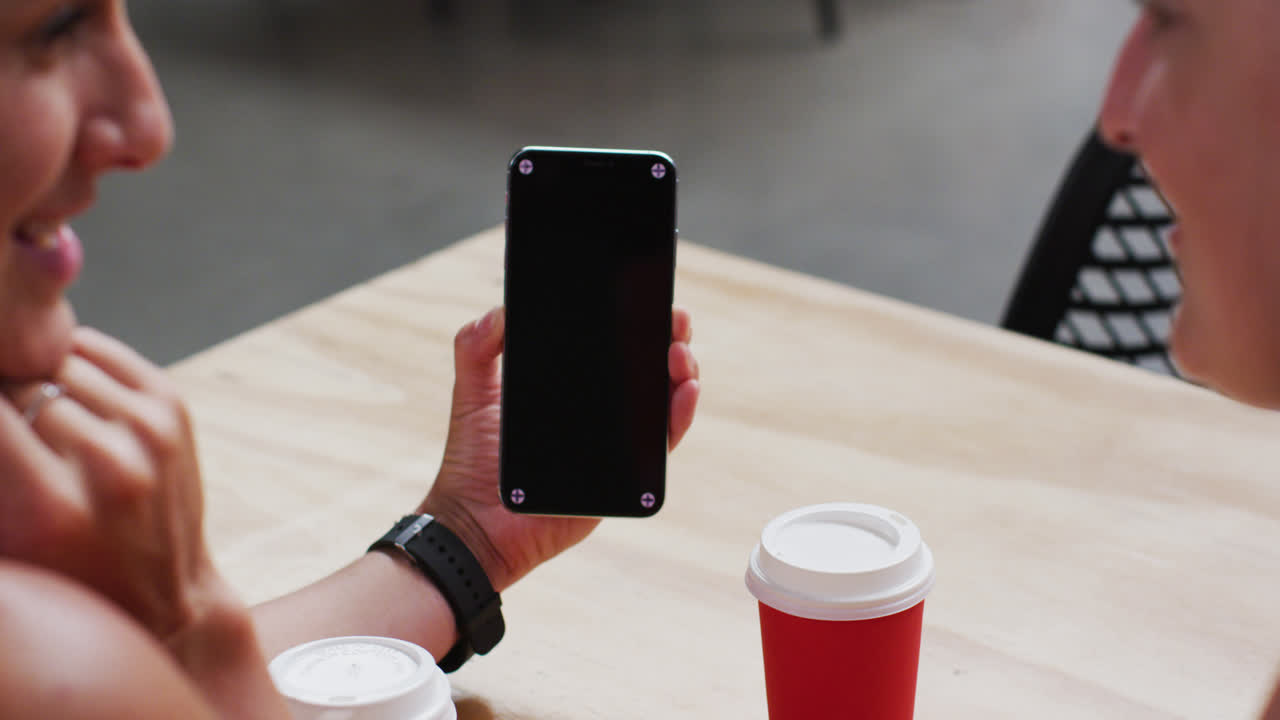 Vista da sopra la spalla di due donne caucasiche felici che guardano lo smartphone al tavolo in un caffè