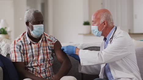 senior caucasian male doctor vaccinating senior african american man at home both wearing face masks