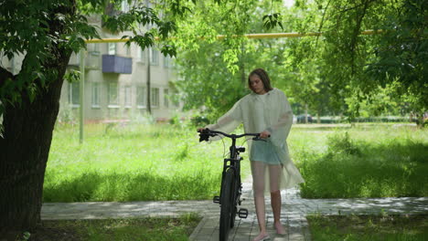 blonde woman in raincoat thoughtfully strolling with bicycle on paved path through green park, surrounded by trees, grass, and urban background with a yellow pole