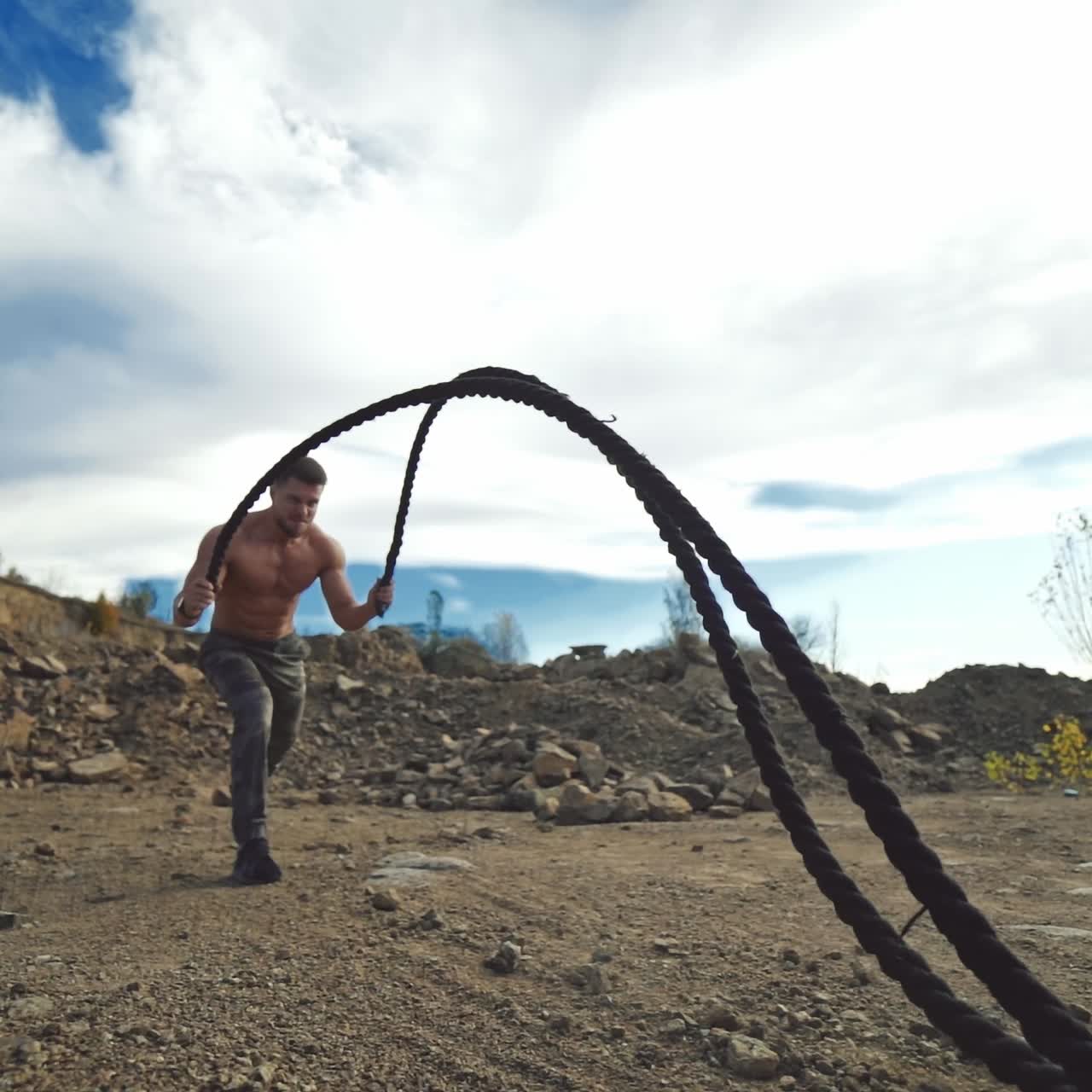 Professional sportsman working out with battle ropes. Muscular man without shirt pumping muscles with cables on the nature rocky background.