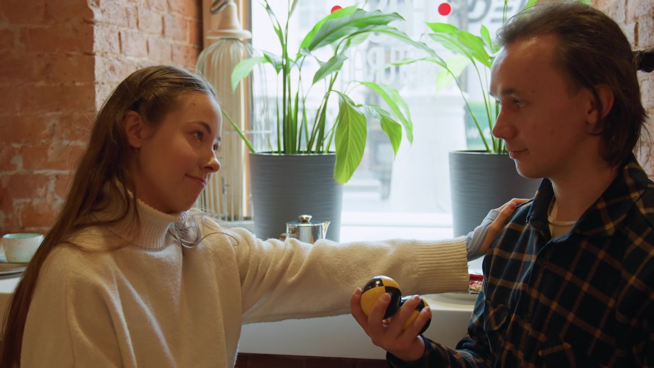 Close up of two people interacting in cozy cafe setting one holding black and yellow balls while other engages. plate with food items like sandwich and cup sits on counter near plants