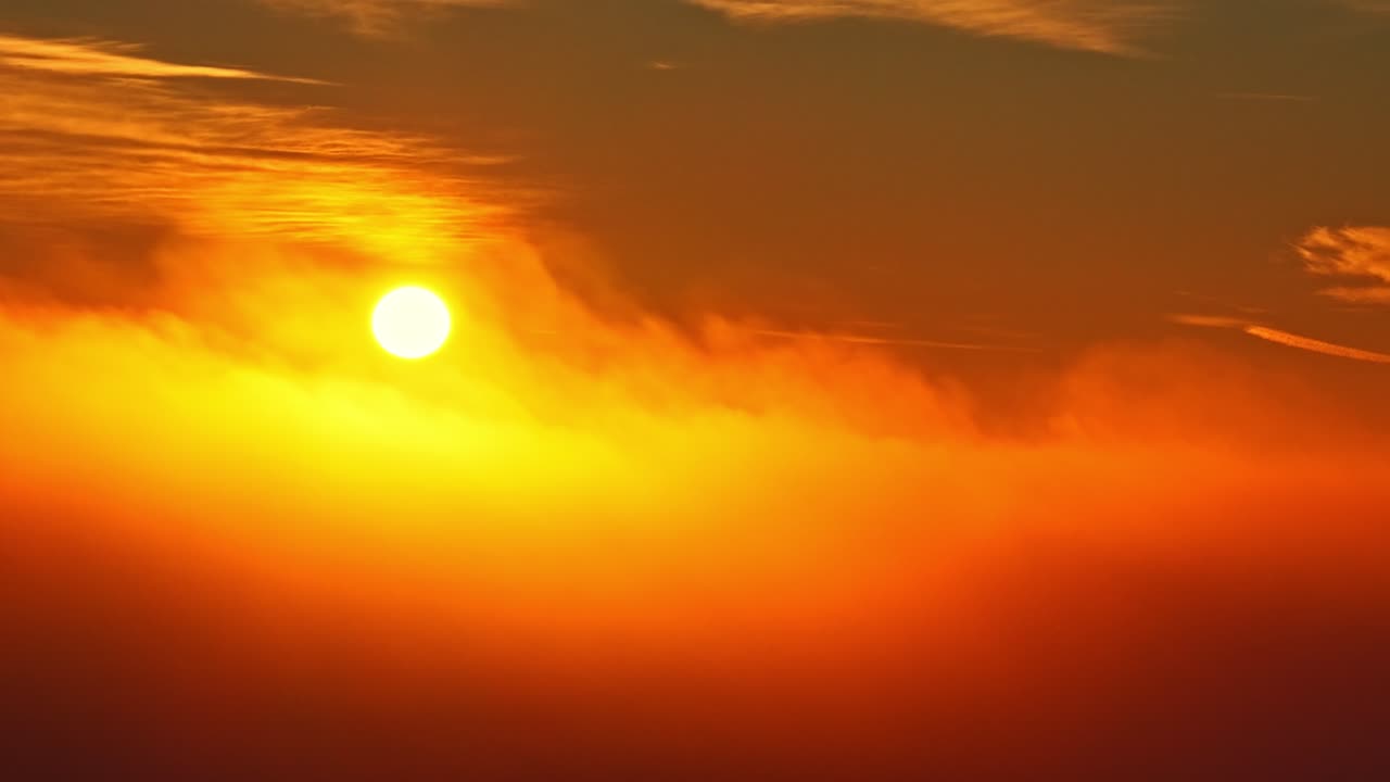 Drone view of the sun rising or setting above soft clouds, glowing in warm orange and yellow tones. Gentle sunlight along the horizon creates a calm, uplifting background with a peaceful mood