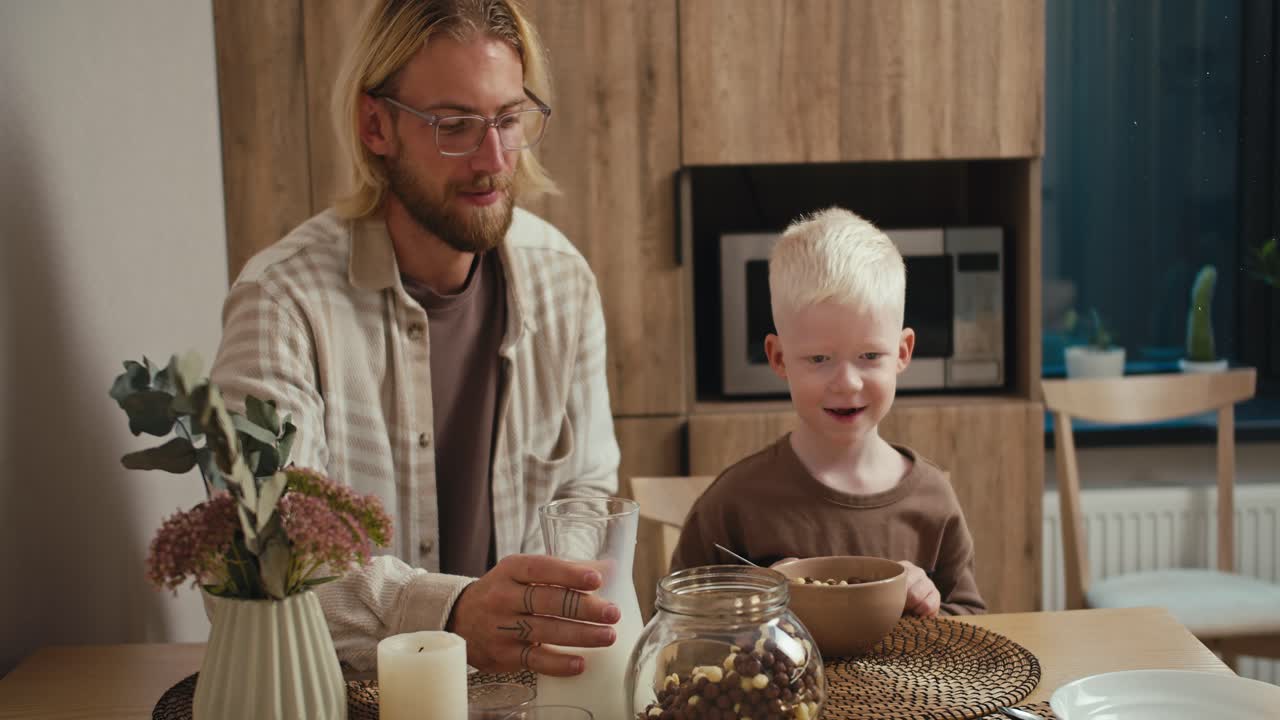 A blond man with glasses in a white checkered shirt pours milk into the bowl of his little albino son with white hair color. And the boy eats his breakfast in the morning in a modern kitchen