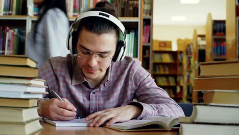 young caucasian male student with big headphones is sitting at table in library. many books are aroung him, he is smiling listening to music and writing